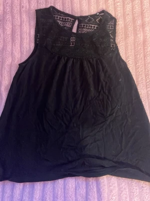 Svart ärmlös topp med spetsdetaljer - Snygg svart ärmlös topp med spetsdetaljer upptill och rund halsringning. Toppen har en lätt A-linjeform och ett litet nyckelhål bak i nacken. Perfekt att styla med jeans eller kjol för en trendig look.