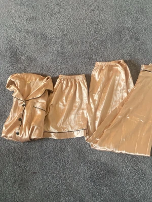 Beige satinpyjamas set med shorts - Snyggt pyjamasset i beige satin med svart kantband. Setet består av en kortärmad skjorta med knappar, ett par shorts och två olika långa byxor, alla med resår i midjan. Perfekt för chillkvällar hemma och ger en lyxig känsla.