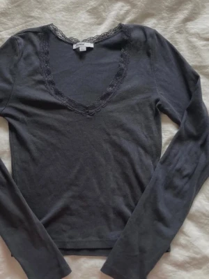 Mörkgrå långärmad topp med spets - Mysig mörkgrå långärmad topp med v-ringning och spetsdetaljer runt halsen. Perfekt för lager på lager och enkel att styla till olika looks. Tunn och mjuk i materialet, passar dig som gillar en enkel men ändå snygg stil.