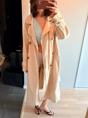 Beige trenchcoat med knytskärp - Stilren beige trenchcoat med dubbelknäppning, bred krage och knytskärp i midjan. Jackan har stora fickor och klassiska detaljer vid ärmsluten. Perfekt för lager på lager och ger en cool, avslappnad vibe. Materialet ser ut att vara bomull eller bomullsblandning.
