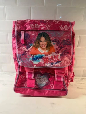 Rosa Violetta ryggsäck med motiv - Säljer en rosa ryggsäck med Violetta-motiv och texten 'Love Music Passion'. Väskan har flera fack, justerbara axelband och detaljer i vitt och lila. Perfekt för dig som gillar musik och färgglada accessoarer. Tillverkad i slitstark polyester med tryckta mönster och hjärtan.