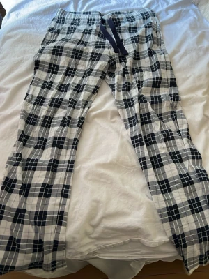 Rutiga pyjamasbyxor i bomull - Säljer ett par klassiska pyjamasbyxor med svartvitt rutmönster och dragsko i midjan. Byxorna är raka i modellen och gjorda i mjuk bomull, perfekta för chill hemma eller sovstunder. Passar dig som gillar enkel och tidlös stil.