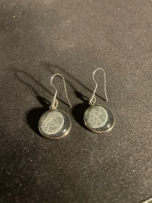 Runda örhängen med grått/blått mönster - Säljer ett par runda örhängen med silverfärgade krokar och en grå/blå, glansig yta med ett abstrakt vitt mönster. Örhängena har en modern och stilren look som passar till många olika stilar. OBS! Materialet är okänt, kan ej garantera att det är rostfritt.