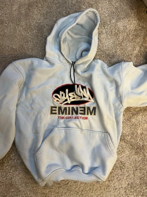 Ljusblå Eminem hoodie med tryck - Säljer en ljusblå hoodie med Eminem-tryck och texten 'The Collection' på bröstet. Hoodien har en stor magficka, dragsko i huvan och coolt broderat motiv. Perfekt för dig som gillar streetwear och vill sticka ut lite extra.