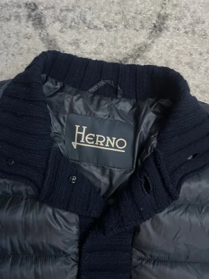 Mörkblå pufferjacka från Herno - Snygg mörkblå pufferjacka från Herno med ribbstickad krage, muddar och knappslå. Jackan har en quiltad design och stängs med knappar framtill. Perfekt för kalla dagar och ger en stilren look.