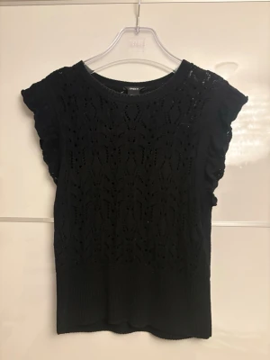 Svart stickad topp med volangärm - Snygg svart stickad topp från Lindex med volangdetaljer vid ärmarna och ett genombrutet mönster över hela plagget. Toppen har rund halsringning och är ärmlös med en feminin touch. Perfekt att styla med jeans eller kjol. Storlek S✨💞