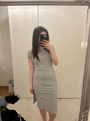 Grå ribbad midiklänning - Snygg grå ribbad klänning med korta ärmar och rund hals. Klänningen har slits på båda sidor och är i en mjuk, stretchig bomullsblandning. Perfekt till sneakers eller boots för en avslappnad look.