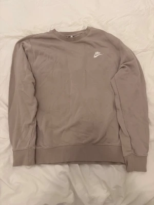 Beige sweatshirt från Nike - En stilren beige sweatshirt från Nike med klassisk rund halsringning och diskret Nike-logga broderad i vitt på bröstet. Tröjan har långa ärmar och ribbade muddar vid ärmslut och nederkant. Perfekt för en avslappnad och sportig look.