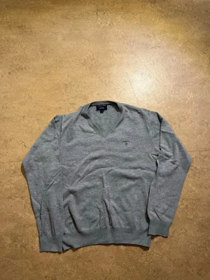 Grå V-ringad tröja från Gant - Klassisk grå V-ringad tröja från Gant med diskret logga broderad på bröstet. Tröjan har långa ärmar, ribbade muddar och är tillverkad i mjukt material som passar perfekt till lager på lager. Enkel och stilren design.