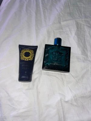 Versace Dylan Blue Eau de Toilette - Versace Dylan Blue Eau de Toilette i en elegant mörkblå fyrkantig glasflaska med gulddetaljer. Medföljer en matchande mörkblå tub med guldmönster, troligen en aftershave balm eller duschgel. Lyxig och modern design som sticker ut i badrumshyllan.