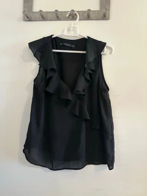 Svart volangblus från Zara - Snygg svart blus från Zara med volangdetaljer längs ringningen. Ärmlös modell i ett lätt och luftigt material som ger en elegant look. Perfekt att styla med jeans eller kjol för en trendig outfit.
