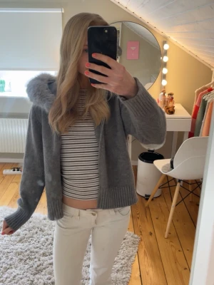 Grå tunn jacka med fluffig luva  - Mysig grå hoodie med dragkedja och stor luva kantad med fluffig fuskpäls. Tröjan har ribbade muddar och är gjord i ett mjukt stickat material. Storleken står inte men jag gissar att den är i S!