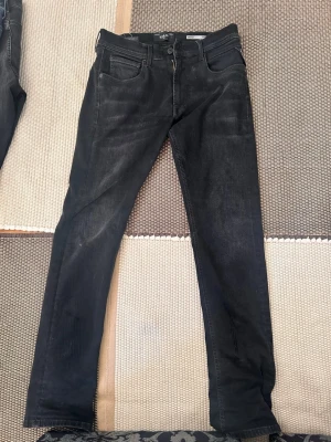 Svarta slim fit jeans från Nudie - Säljer ett par svarta slim fit jeans från Replay Jeans med klassisk femficksmodell och subtila slitningar. Jeansen har raka ben och är tillverkade i mjuk denim med stretch för extra komfort. Perfekta för en avslappnad och snygg stil.