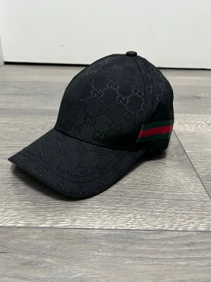 Svart Gucci keps med GG-mönster - Snygg svart keps från Gucci med klassiskt GG-monogram över hela kepsen. På sidan finns den ikoniska gröna och röda randen. Kepsen har böjd skärm och justerbar rem bak. Materialet är slitstarkt tyg som ger en exklusiv känsla.