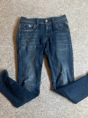 Mörkblå Replay jeans med slitningar - Säljer ett par mörkblå jeans från Replay med snygga slitningar och tvättade detaljer. Klassisk femficksmodell med knappgylf och Replay-logga vid fickan. Perfekta för en avslappnad och trendig look.