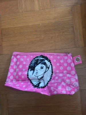 Rosa necessär med prickar och porträtt - Söt rosa necessär med vita prickar och ett svartvitt porträtt av en tjej på framsidan. Väskan har dragkedja upptill och en metallring i hörnet. Tillverkad i plastmaterial, perfekt för smink eller småprylar.