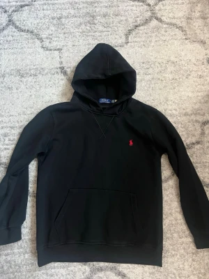 Svart hoodie från Polo Ralph Lauren - Snygg svart hoodie från Polo Ralph Lauren med klassisk röd logga broderad på bröstet. Hoodien har huva, känguruficka och ribbade muddar. Tillverkad i mjukt bomullsmaterial som är skönt att bära. Perfekt för en avslappnad och stilren look.