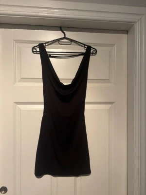 Svart kort klänning med draperad urringning - Säljer en svart kort klänning med snygg draperad urringning och breda axelband. Klänningen har en figurnära passform och är perfekt för dig som gillar en stilren och enkel look. Materialet känns mjukt och stretchigt, troligen polyester eller syntet.