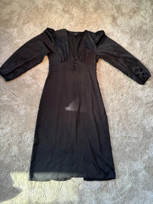 Svart långklänning från Gina Tricot - Svart midiklänning från Gina Tricot med v-ringning, puffiga långärmar och dekorativa knappar framtill. Klänningen har en elegant, figurnära passform och är tillverkad i ett lätt, glansigt material som ger en lyxig känsla.   Klänningen går till mitten av vaderna ungefär  Har ett litet hål under armen, inget som syns jättemycket när man har på sig den.