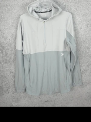 Ljusgrå hoodie/jacka från Under Armour - Snygg ljusgrå hoodie från Under Armour med dragkedja vid halsen och stor huva. Tillverkad i ett mjukt funktionsmaterial som andas. Har långa ärmar med diskret logga på mudden och Under Armour-logga på bröstet. Perfekt för träning eller chill.