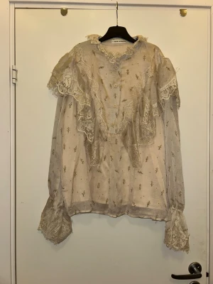 Romantisk spetsblus från Sofie Schnoor  - Drömmig beige blus från Zara med romantiska spetsdetaljer och volanger. Blusen har små broderade blommor, knappar framtill och hög krage med spets. Långa ärmar med breda spetsmanschetter ger en vintagekänsla. Perfekt för dig som gillar en feminin och unik stil.