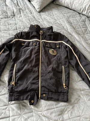 Svart vindjacka från Nickelson - Snygg svart vindjacka från Nickelson med guldfärgade dragkedjor och detaljer. Jackan har två bröstfickor med knappar, två sidofickor med dragkedja och ett stort emblem på bröstet med texten 'Nickelson Established 1954'. Materialet är glansigt och lätt.