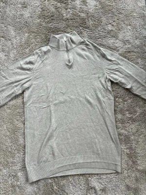 Ljusgrå stickad tröja med halv dragkedja - Ljusgrå stickad tröja med hög krage och halv dragkedja framtill. Tröjan har långa ärmar och ribbade muddar vid ärmslut och nederkant. Perfekt för lager på lager och enkel att matcha med olika outfits. OBS! Barnstorlek 176.