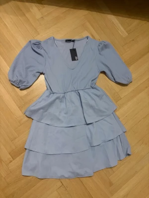 Ljusblå volangklänning med v-ringning - Supersöt ljusblå klänning med v-ringning, puffiga trekvartsärmar och flera volanglager i kjolen. Klänningen har en feminin siluett och resår i midjan för en bekväm passform. Perfekt för vår och sommar!