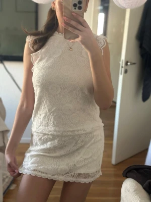 Vit spetsklänning med ärmlös design - Säljer en söt vit kortklänning i spets med rund halsringning och ärmlös design. Klänningen har fina spetsdetaljer och en figurnära passform som ger en romantisk look. Perfekt för sommarens alla tillfällen!