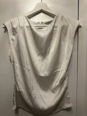 Vit ärmlös sidenblus - Säljer en elegant vit blus i siden med draperad front och ärmlös design. Perfekt för dig som gillar en stilren och lyxig look. Blusen har en mjuk och glansig yta som ger en exklusiv känsla.