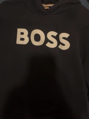 Marinblå hoodie från BOSS - Snygg marinblå hoodie från BOSS med stor vit logga på bröstet. Klassisk modell med huva och ribbade muddar vid ärmslut och nederkant. Perfekt för en avslappnad och trendig stil. Materialet känns mjukt och bekvämt.