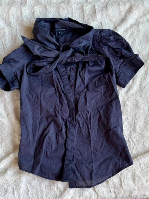 Blå blus med rosett i kragen - Snygg blå blus från french connection med puffiga korta ärmar och en stor rosett i kragen. Blusen har knappar framtill och är gjord i ett tunt, vävt material som känns lätt och luftigt. Perfekt för dig som vill ha en trendig och unik look.