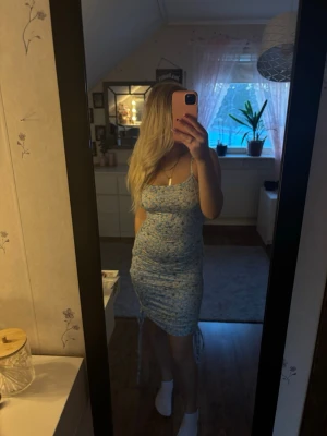 Blommig blå klänning - Söt blå kortklänning med blommigt mönster och smala axelband. Klänningen har en åtsittande passform och snörningsdetaljer på sidorna som ger en snygg siluett. Perfekt för varma dagar och somriga tillfällen. Man kan skystera själv hur lång eller kort man vill ha med hjälp av snörerna🌺☀️