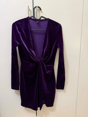 Lila sammetsklänning med knyt - Snygg lila sammetsklänning med lång ärm och djup v-ringning. Klänningen har ett knyt framtill som ger en cool twist och markerar midjan. Kort modell som passar perfekt till säsongens alla tillfällen. Perfekt för dig som vill sticka ut!