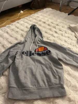 Grå Ellesse hoodie med logga - Säljer en grå hoodie från Ellesse med stor logga i marinblått, orange och rött på bröstet. Hoodien har huva med vita snören och en stor magficka framtill. Tillverkad i mjukt sweatshirtmaterial och har ribbade muddar vid ärmar och nederkant.