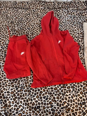 Nike tech fleece röd  - Säljer ett rött loungewear set från Nike med matchande hoodie och byxor. Hoodien har dragkedja, fickor och vit Nike-logga på bröstet. Byxorna har resår i midjan och samma logga på benet. Perfekt för chill dagar hemma.