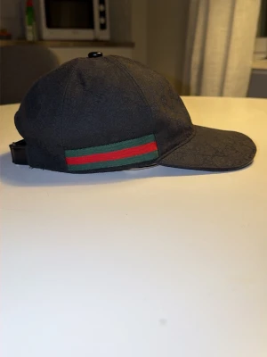 Svart keps från Gucci med grön/röd detalj - Säljer en svart keps från Gucci med klassiskt grönt och rött band på sidan. Kepsen har böjd skärm och justerbart spänne i svart läder baktill. Materialet är tyg med diskret GG-mönster som ger en exklusiv känsla. Perfekt för dig som gillar streetwear och lyxiga accessoarer. Pris kan diskuteras vid snabb o enkel affär