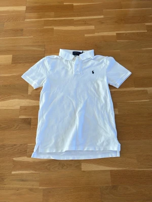 Vit pikétröja från Ralph Lauren - Klassisk vit pikétröja från Ralph Lauren med korta ärmar och krage. Tröjan har knappar framtill och den ikoniska lilla logotypen broderad på bröstet. Tillverkad i mjuk bomull som känns skön mot huden. Perfekt till jeans eller shorts.