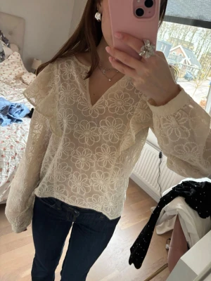 Vit blommig spetsblus med volanger - Söt vit blus i transparent spets med blommigt mönster och volangdetaljer längs axlarna. V-ringad hals och långa ärmar med resår vid ärmslut. Perfekt för en romantisk look och enkel att styla till jeans eller kjol.