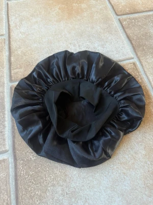 Svart satin hårbonnet - Snygg svart hårbonnet i glansigt satinmaterial. Perfekt för att skydda håret under natten och minska friss. Bonneten har en elastisk kant som gör att den sitter bekvämt på huvudet. Enkel och stilren design som passar alla hårtyper.