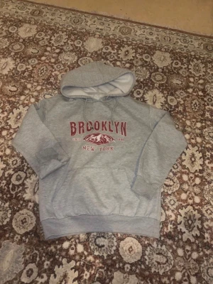Grå hoodie med Brooklyn-tryck - Säljer en grå hoodie med huva och dragsnören. På bröstet står det 'BROOKLYN NEW YORK' i vinrött tryck tillsammans med ett bergmotiv och texten 'Since 1898'. Hoodien har magficka och ribbade muddar. Perfekt för en chill och avslappnad stil.