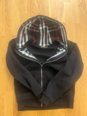 Svart hoodie med rutigt foder - Säljer en svart hoodie från Burberry med dragkedja och klassiskt rutigt foder i huvan. Hoodien har fickor framtill och ribbade muddar vid ärmslut och nederkant. Perfekt för dig som gillar stilrena detaljer och ikoniskt mönster.Dragkedjan är lite sönder men går och fixas annars har den inga håll eller nått 