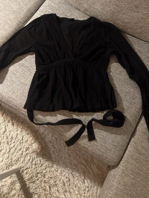 Svart omlottopp med knytband - Snygg svart omlottopp med långa ärmar och knytband i midjan. Toppen har en v-ringad urringning och är tillverkad i ett mjukt, lätt material som ger en skön passform. Perfekt för att skapa en stilren look.