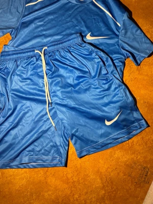 Blått Nike träningsset - Säljer ett blått träningsset från Nike med matchande t-shirt och shorts. Setet har vita Nike-loggor och är gjort i ett lätt, snabbtorkande material som passar perfekt för träning eller fotboll. Shortsen har dragsko i midjan för bästa passform.