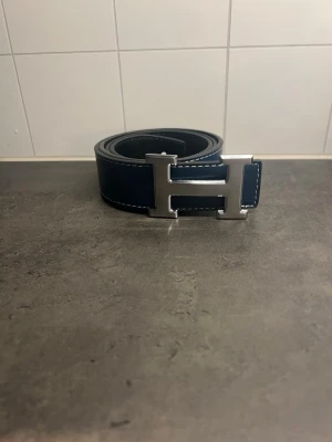 Herme bälte - Snyggt blått bälte med ett stort H-format spänne i silverfärg. Skärpet har vita sömmar längs kanterna som ger en clean kontrast. Perfekt accessoar för att lyfta din outfit.