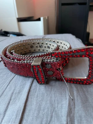 Rött glittrigt bälte med nitar - Säljer ett riktigt statement-bälte i rött med glittriga detaljer och silverfärgade nitar. Spännet är stort och dekorerat med röda stenar som matchar bältets glittriga yta. Perfekt för att lyfta vilken outfit som helst.