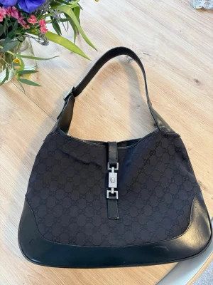 Gucci Jackie Bag - Snygg svart axelremsväska från Gucci i modellen vintage Jackie, med klassiskt GG-mönster i tyg och detaljer i svart läder. Väskan har en justerbar rem och silverfärgat spänne framtill. Medföljer matchande dustbag i samma GG-tyg. Perfekt för dig som gillar lyxiga accessoarer. SIZE MEDIUM 22x32x5. Väskan är i bra skick men defekter på läder remen finns (se bilder), det går att åtgärda hos en lädermakare eller skomakare.  Äkthetsbevis finns💓 pris går att diskutera 