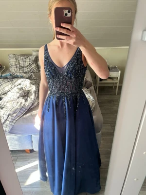 Marinblå långklänning med paljetter - Säljer en marinblå långklänning med v-ringning och smala axelband. Överdelen är dekorerad med glittriga paljetter och har en elegant, figurnära passform medan kjolen faller i ett mjukt, svepande tyg. Perfekt för bal eller festkväll!