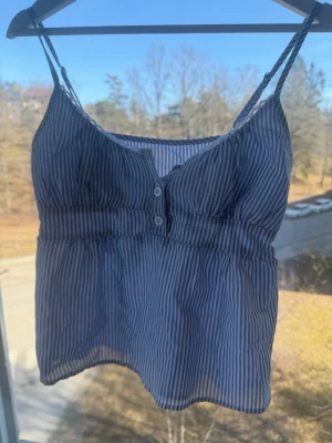 Blåvit randig linnetopp - Snygg blåvit randig linnetopp med tunna axelband och två knappar framtill. Toppen har en lätt rynkad midja och är tillverkad i ett tunt, luftigt bomullsmaterial. Perfekt för varma dagar och enkel att styla med jeans eller kjol. lappen är borta, så kan icke se vad märken eller stoleken är men skulle tippa på s! 