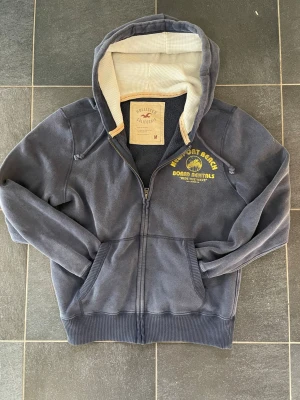 Blå Hollister hoodie - Blå hoodie från Hollister med dragkedja och huva. På bröstet och ryggen finns gult tryck med texten 'Newport Beach Board Rentals' och palmmotiv. Insidan av huvan är ljus och ribbade muddar finns vid ärmslut och nederkant. Perfekt för en avslappnad outfit till våren och sommaren! Storlek M i nytt skick! 